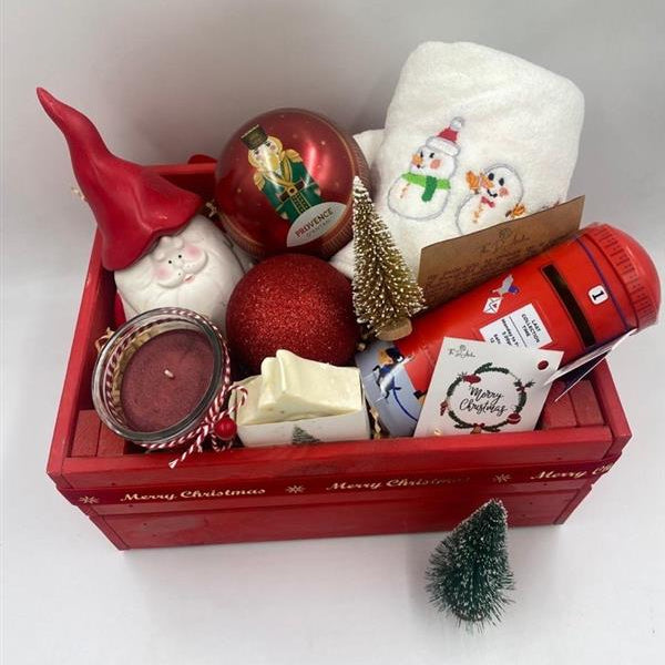 A Wooden festive hamper, red color, having Christmas towel, a ball Christmas ornament with tea bags inside, candle, porcelain Santa's container, natural handmade festive soap bar with Christmas tag, Christmas chocolate