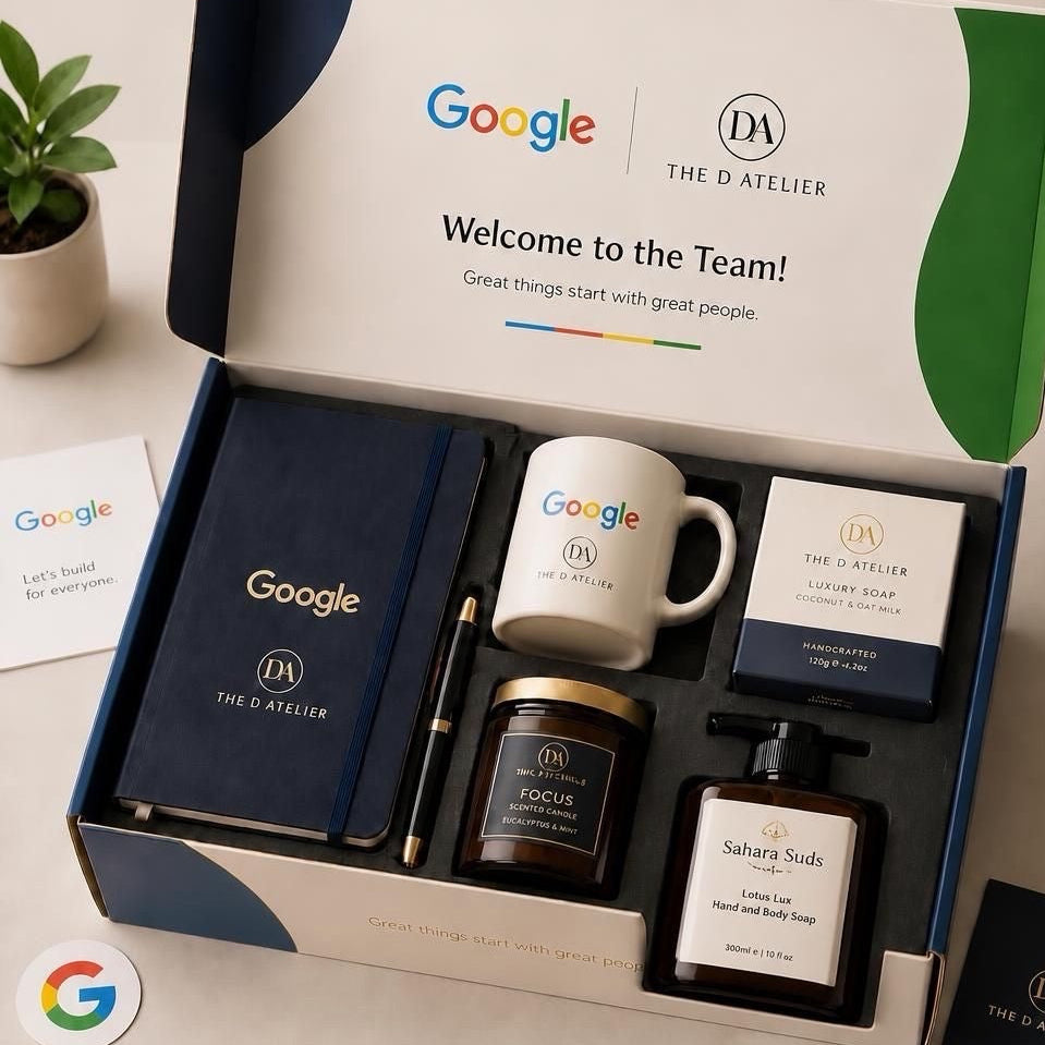 A Google and The D Atelier team gift set displayed in an open branded box on a light surface. The set includes a navy notebook, a white mug with Google and DA logos, a candle jar labeled Focus, a soap box labeled Luxury Soap, and a hand and head soap bottle labeled Sahara Suds.