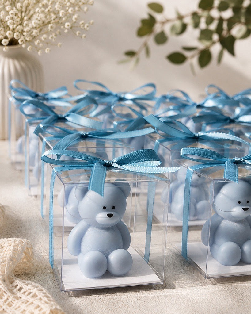 A row of small clear favor boxes tied with light blue ribbons, each containing a pale blue teddy bear figurine, arranged on a soft beige surface with decorative flowers and greenery in the background.