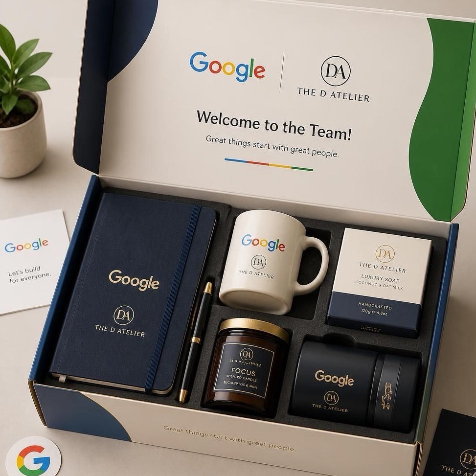 A branded Google x The D'Atelier gift set displayed in an open box on a light beige surface. The set includes a navy notebook, a white mug with Google and The D'Atelier branding, a candle tin, a small box, and two black Google-branded items.