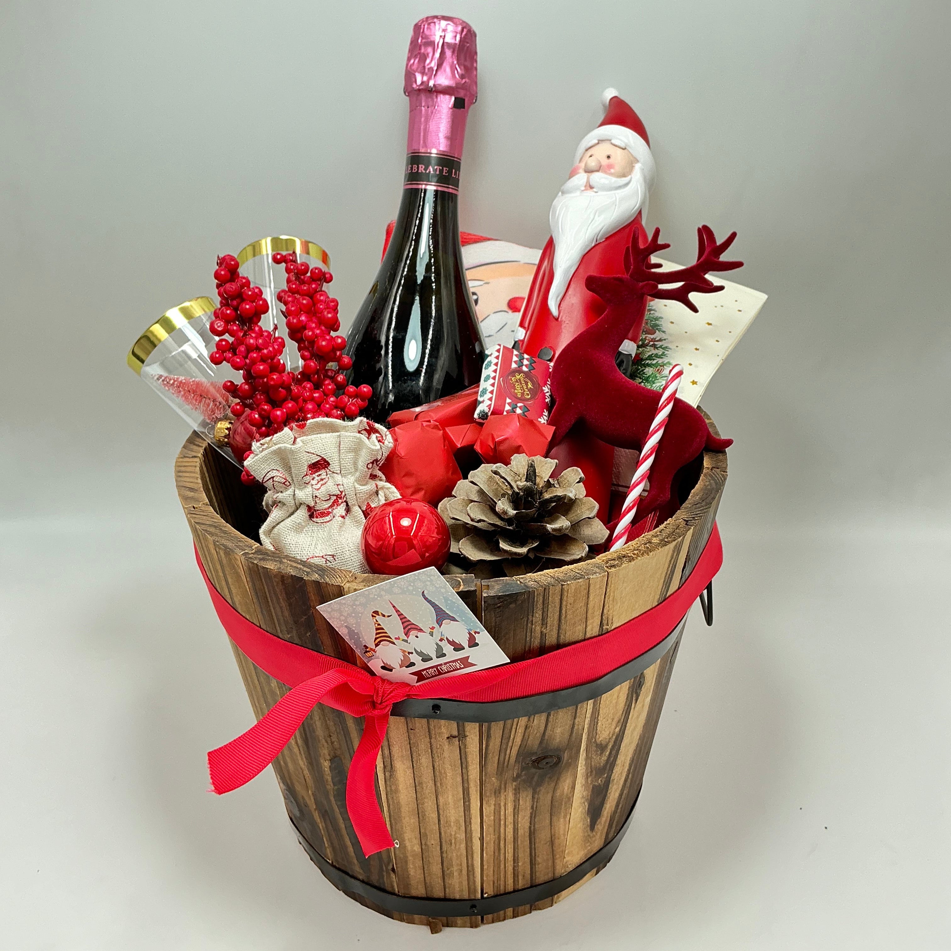 A gift basket featuring a Santa Claus figure, a velvet reindeer, sparkling non-alcoholic champagne, festive chocolates, a pine cone, two champagne flutes, a jute bag, Christmas ornaments, kitchen towels, and two festive Christmas napkins.