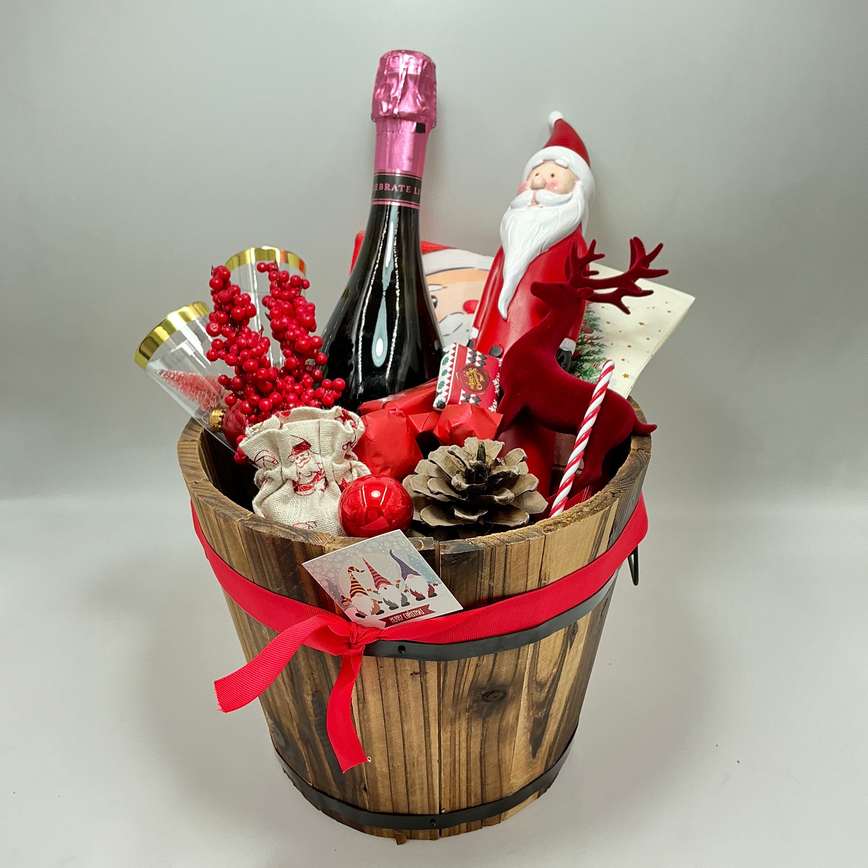 A gift basket featuring a Santa Claus figure, a velvet reindeer, sparkling non-alcoholic champagne, festive chocolates, a pine cone, two champagne flutes, a jute bag, Christmas ornaments, kitchen towels, and two festive Christmas napkins.