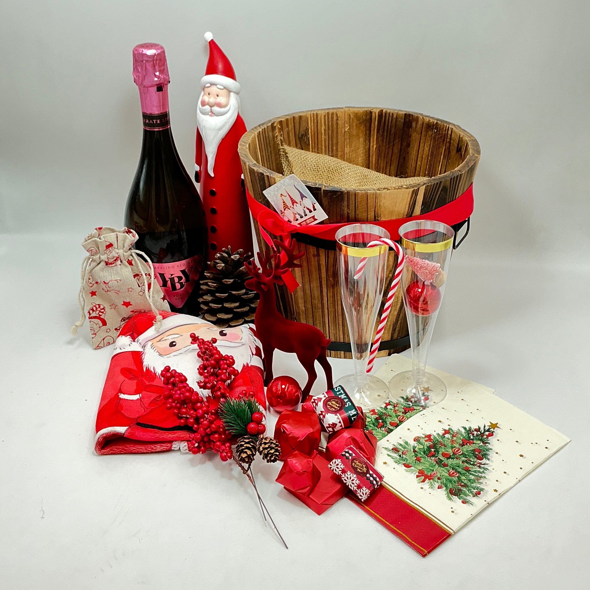 A gift basket featuring a Santa Claus figure, a velvet reindeer, sparkling non-alcoholic champagne, festive chocolates, a pine cone, two champagne flutes, a jute bag, Christmas ornaments, kitchen towels, and two festive Christmas napkins.