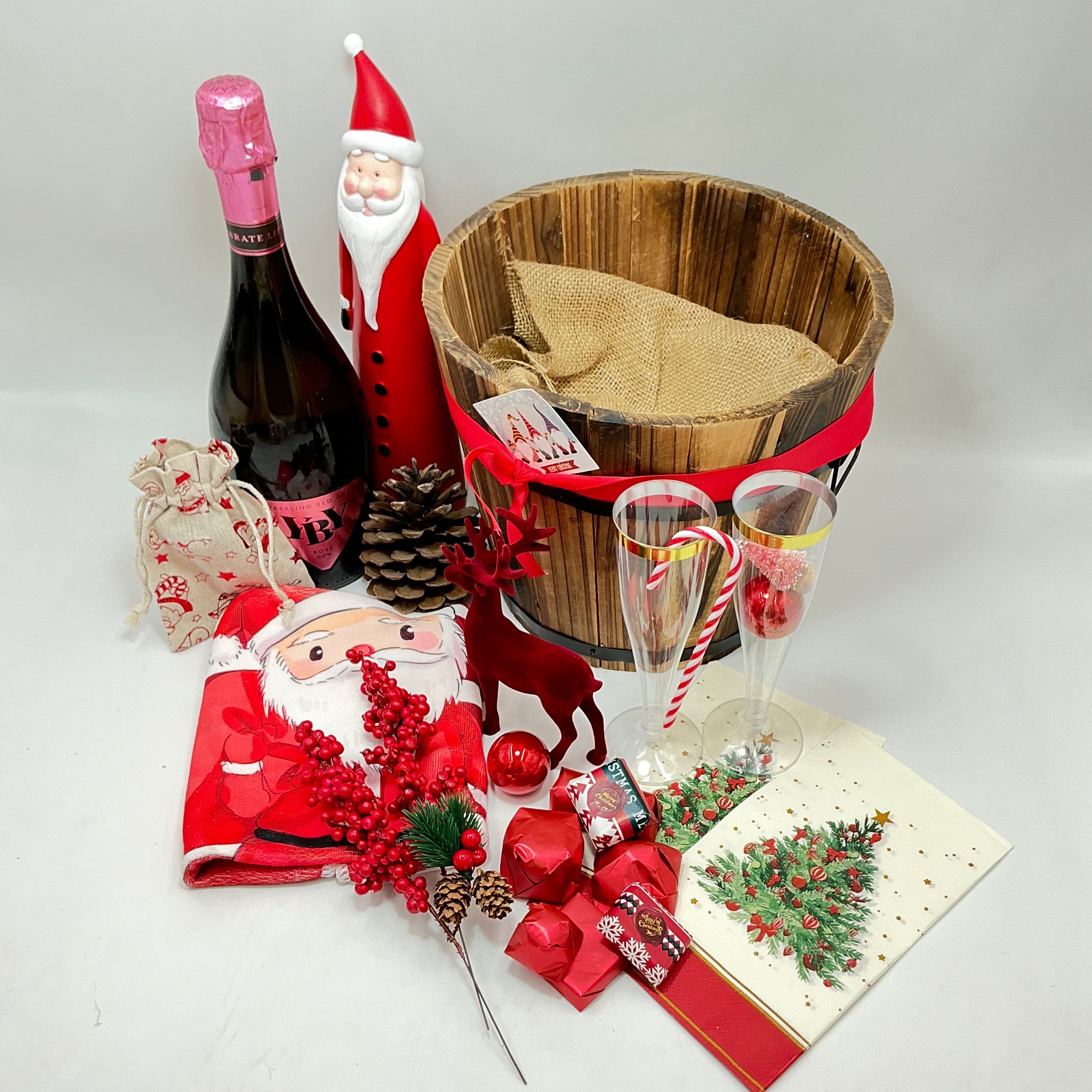 A gift basket featuring a Santa Claus figure, a velvet reindeer, sparkling non-alcoholic champagne, festive chocolates, a pine cone, two champagne flutes, a jute bag, Christmas ornaments, kitchen towels, and two festive Christmas napkins.