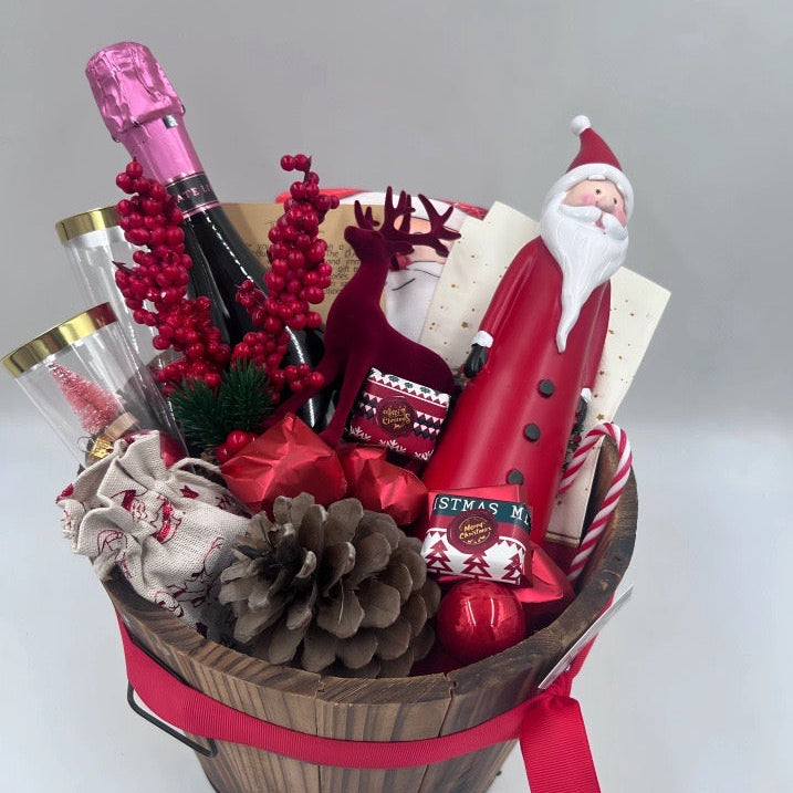 A gift basket featuring a Santa Claus figure, a velvet reindeer, sparkling non-alcoholic champagne, festive chocolates, a pine cone, two champagne flutes, a jute bag, Christmas ornaments, kitchen towels, and two festive Christmas napkins.