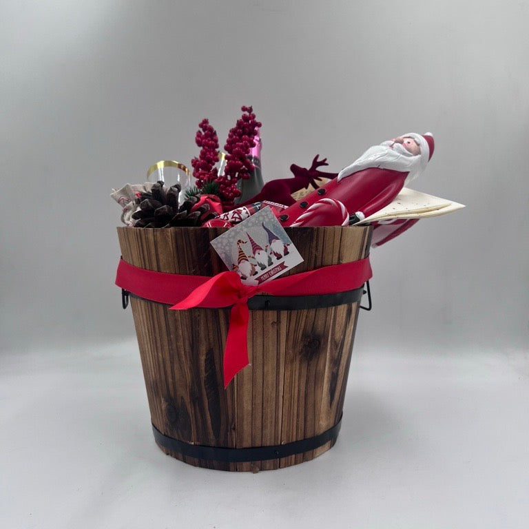 A gift basket featuring a Santa Claus figure, a velvet reindeer, sparkling non-alcoholic champagne, festive chocolates, a pine cone, two champagne flutes, a jute bag, Christmas ornaments, kitchen towels, and two festive Christmas napkins.