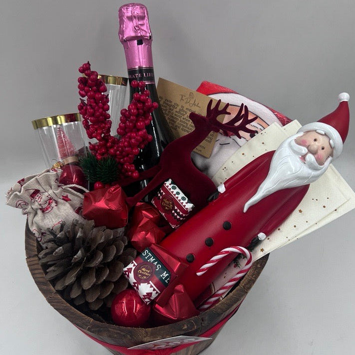 A gift basket featuring a Santa Claus figure, a velvet reindeer, sparkling non-alcoholic champagne, festive chocolates, a pine cone, two champagne flutes, a jute bag, Christmas ornaments, kitchen towels, and two festive Christmas napkins.
