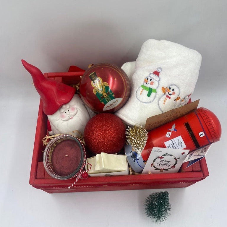 A Wooden festive hamper, red color, having Christmas towel, a ball Christmas ornament with tea bags inside, candle, porcelain Santa's container, natural handmade festive soap bar with Christmas tag, Christmas chocolate  