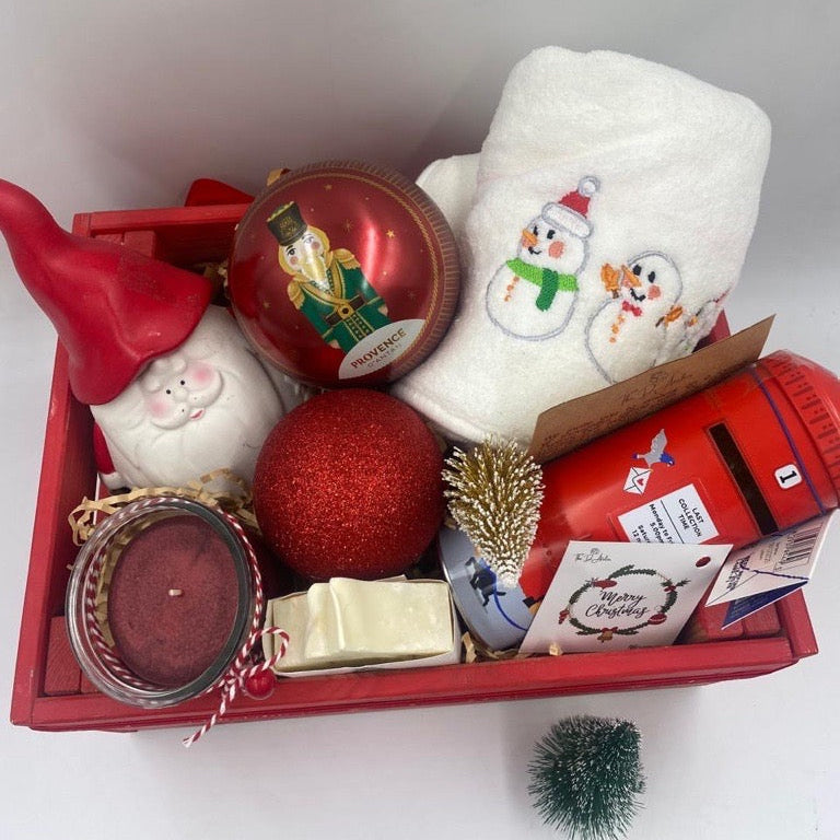 A Wooden festive hamper, red color, having Christmas towel, a ball Christmas ornament with tea bags inside, candle, porcelain Santa's container, natural handmade festive soap bar with Christmas tag, Christmas chocolate  