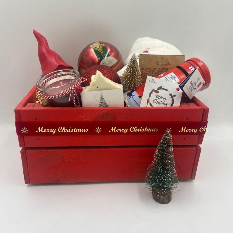 A Wooden festive hamper, red color, having Christmas towel, a ball Christmas ornament with tea bags inside, candle, porcelain Santa's container, natural handmade festive soap bar with Christmas tag, Christmas chocolate  