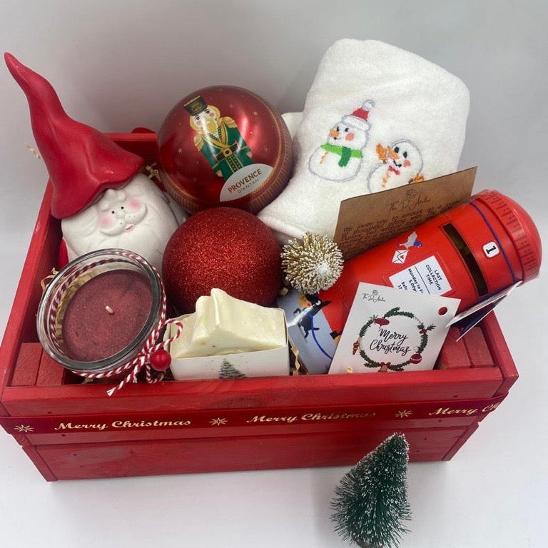 A Wooden festive hamper, red color, having Christmas towel, a ball Christmas ornament with tea bags inside, candle, porcelain Santa's container, natural handmade festive soap bar with Christmas tag, Christmas chocolate  