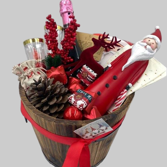 A gift basket featuring a Santa Claus figure, a velvet reindeer, sparkling non-alcoholic champagne, festive chocolates, a pine cone, two champagne flutes, a jute bag, Christmas ornaments, kitchen towels, and two festive Christmas napkins.