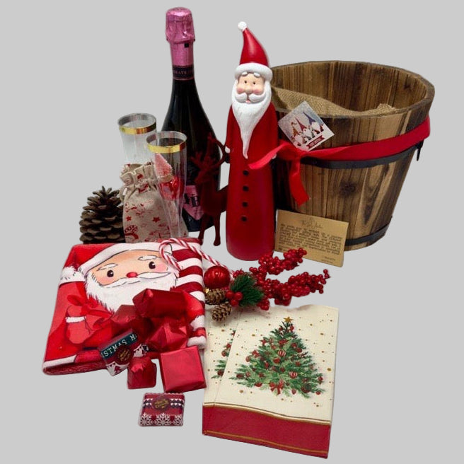 A gift basket featuring a Santa Claus figure, a velvet reindeer, sparkling non-alcoholic champagne, festive chocolates, a pine cone, two champagne flutes, a jute bag, Christmas ornaments, kitchen towels, and two festive Christmas napkins.