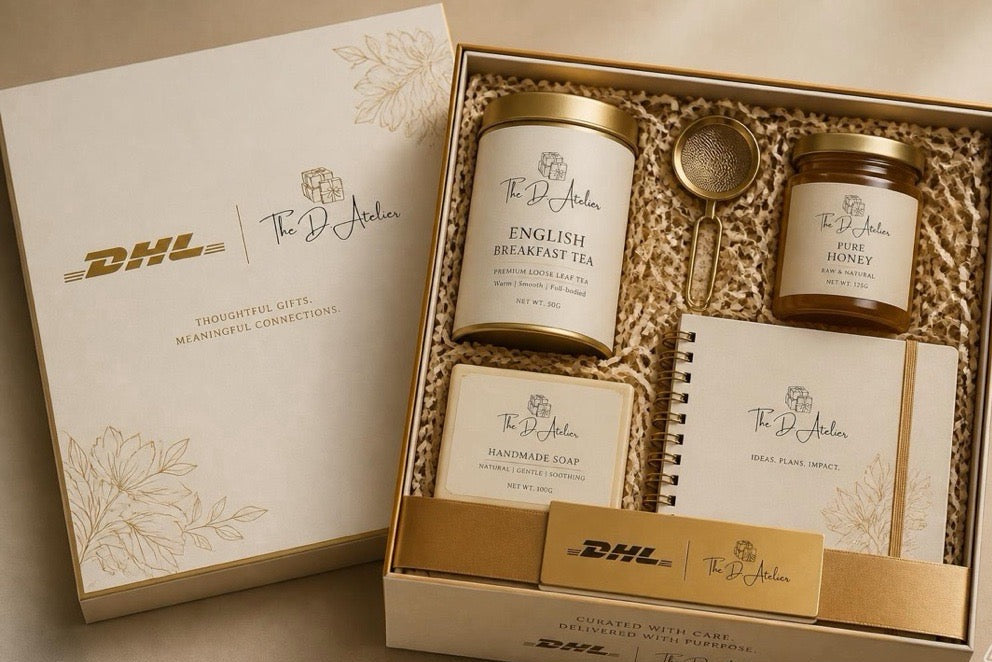 A premium gift set from The D'Atelier Brand, featuring English Breakfast Tea, Honey, Handmade Soap, and a Plane Plate Spoon, presented in a gold and white box with a matching outer box. The DHL brand is prominently displayed on the packaging.