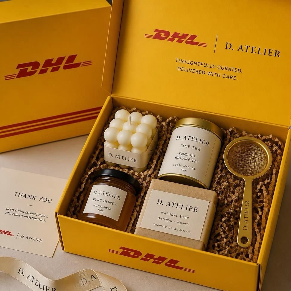 DHL and the DAtelier gift set in a yellow box, containing a candle, a tin of English Breakfast tea, a jar of dark rum, a bar of natural soap, a small metal strainer, and a ribbon, with DHL branding visible on the box and packaging.