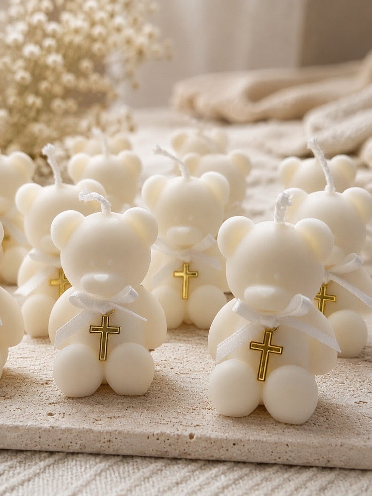 A group of small cream-colored teddy bear candles with gold crosses on their chests, arranged on a textured beige surface with soft, blurred decorative background elements.