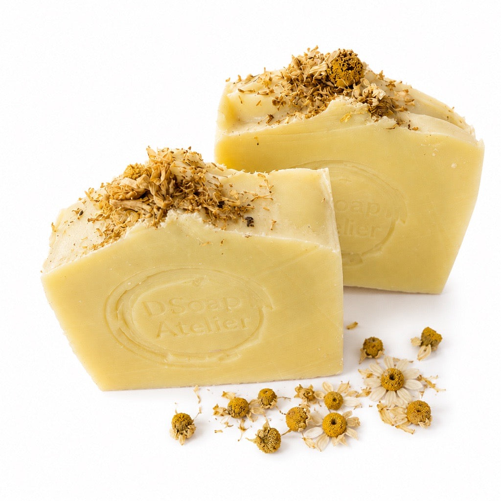 Two bars of pale yellow soap topped with dried chamomile flowers, with a few loose chamomile heads scattered in front, on a white background. The front bar is embossed with the brand name 'DSoap Atelier'.